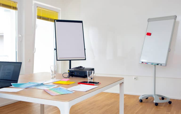 Man sieht einen Raum, in dem Raum steht ein Tisch auf dem Arbeitsmaterial liegt. Im Hintergrund steht ein Whiteboard.
