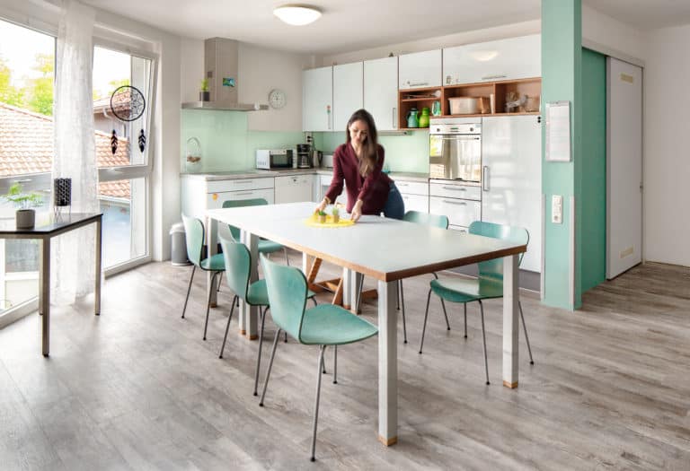 Man sieht einen Wohnraum mit Esstisch und Küche. Eine Frau dekoriert den Tisch.