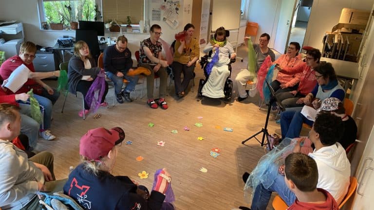 Eine Gruppe von Menschen sitz zusammen in einem Kreis. In der Mitte liegen bunte Papier Blumen.