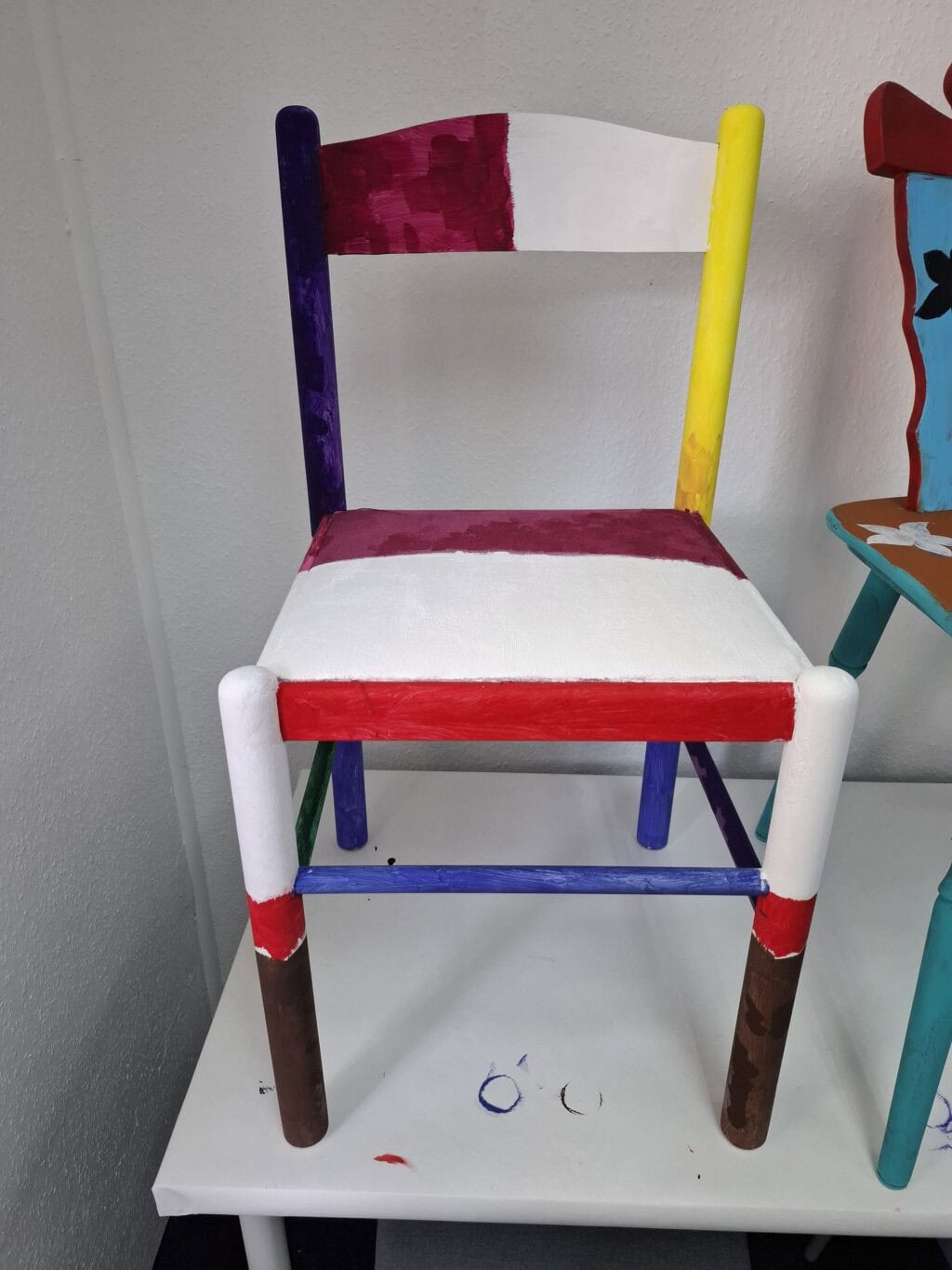Ein bunt bemalter Holzstuhl mit farbigen Flächen in Rot, Lila, Gelb und Weiß.