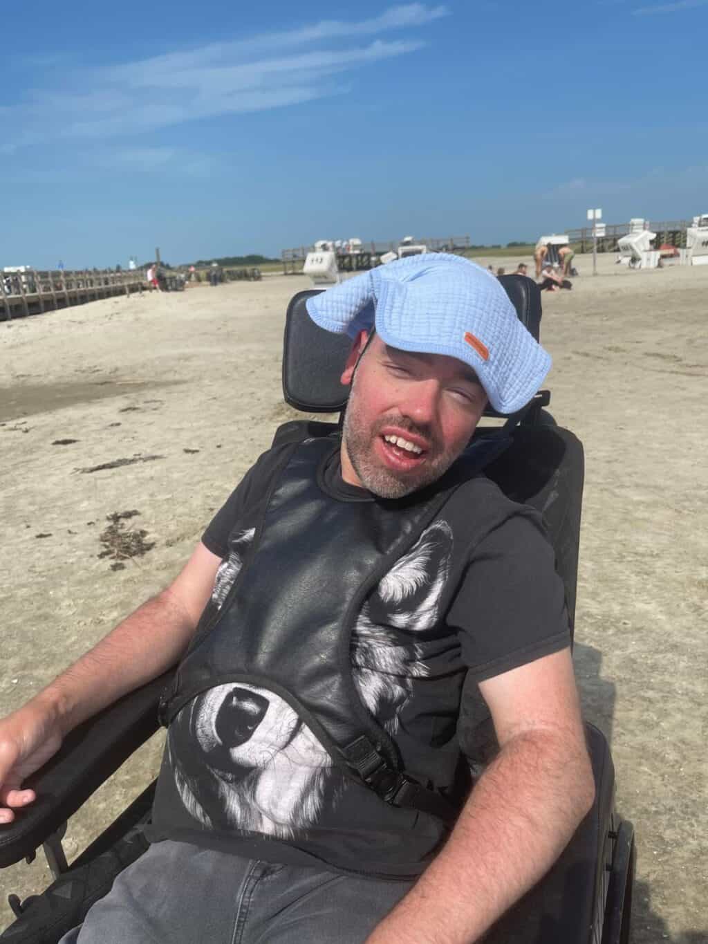 Ein Mann sitzt im Rollstuhl am Strand und lächelt in die Kamera.