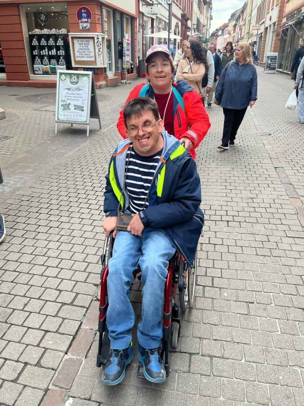 Eine Person schiebt eine andere Person im Rollstuhl durch eine Einkaufsstraße.