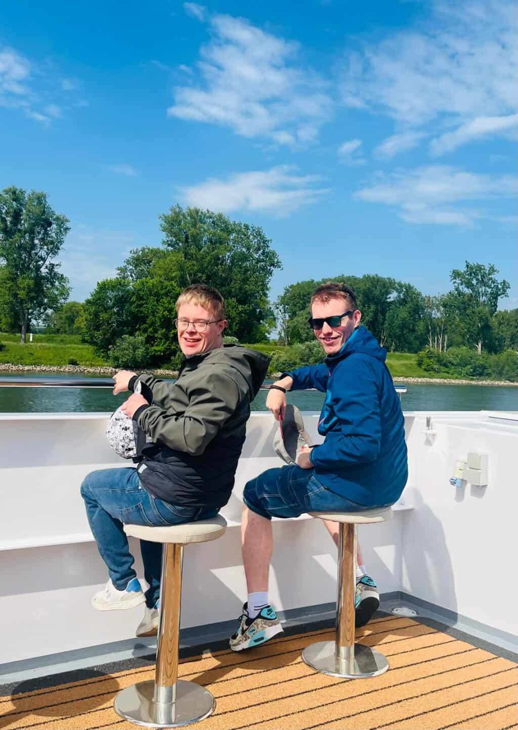 Zwei Männer sitzen auf Barhockern auf einem Schiff und blicken zum Ufer.