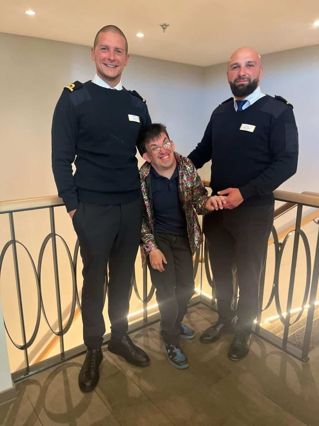 Ein Mann steht lachend zwischen zwei uniformierten Mitarbeitenden des Bootspersonals in einem hellen Innenbereich.