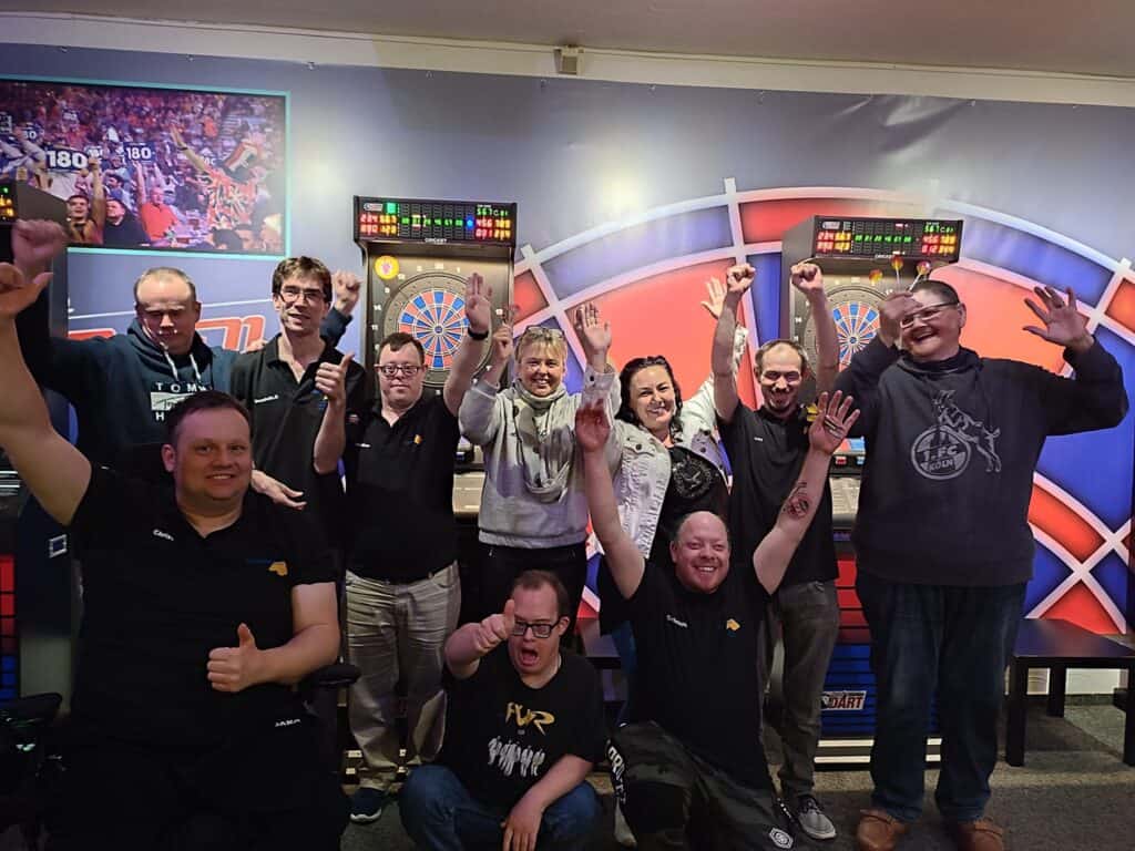 Dart-Team jubelt bei einem Wettbewerb, im Hintergrund Dartboards und eine Leinwand mit Live-Bildern der Veranstaltung.