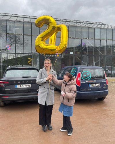 Zwei Personen stehen vor 2 Autos und halten Luftballons in der Hand die eine 20 darstellen.